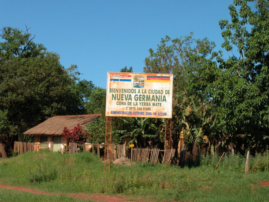 Willkommensschild „Bienvenidos a la ciudad de Nueva Germania“ in Paraguay – Symbol der deutschen Kolonie Nueva Germania mit einfachem Haus und tropischer Umgebung.