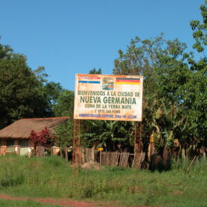 Willkommensschild „Bienvenidos a la ciudad de Nueva Germania“ in Paraguay – Symbol der deutschen Kolonie Nueva Germania mit einfachem Haus und tropischer Umgebung.